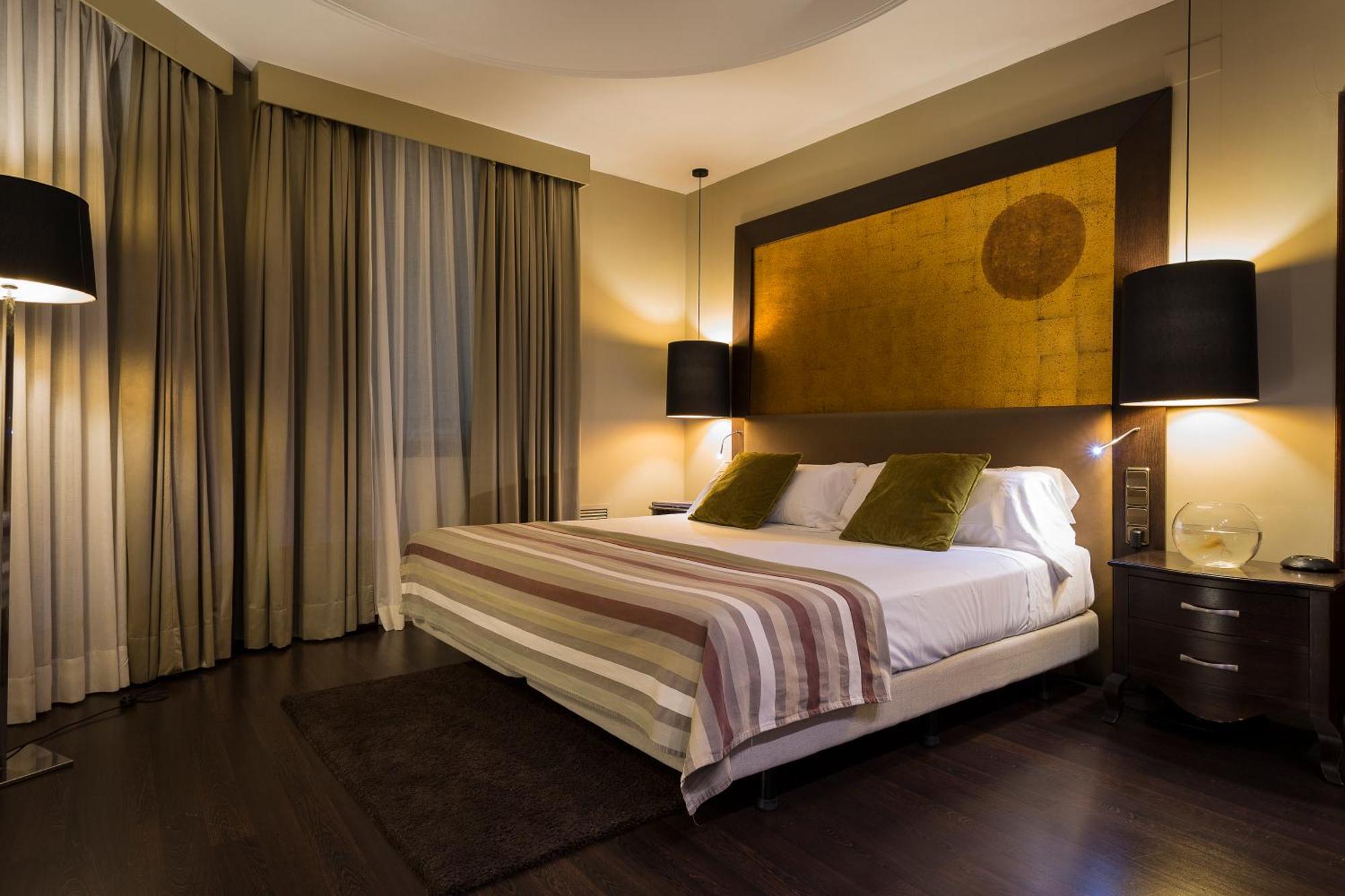 Only You Hotel Valenza Esterno foto The photo shows a modern bedroom that features a large bed with two green accent pillows and a striped bedspread. The room has a warm, neutral color scheme, highlighted by soft lighting from two bedside lamps. The wall behind the bed has an artistic 